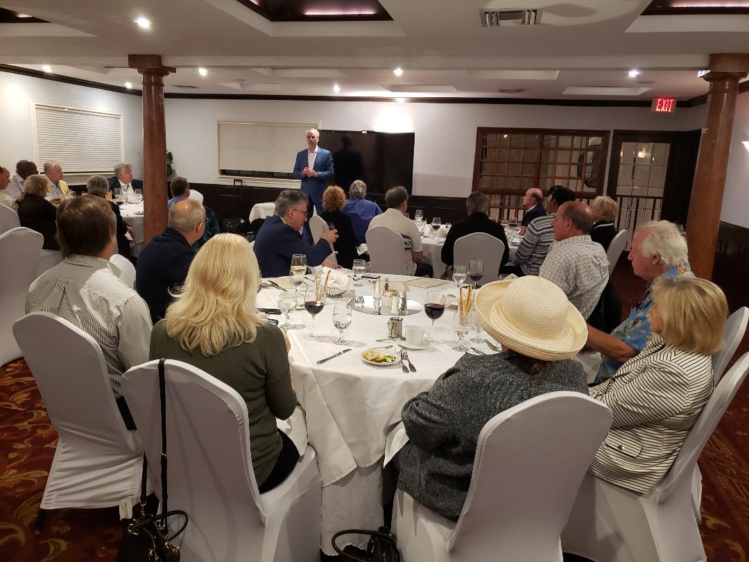 A presenter standing before guests seated at round tables in a banquet room