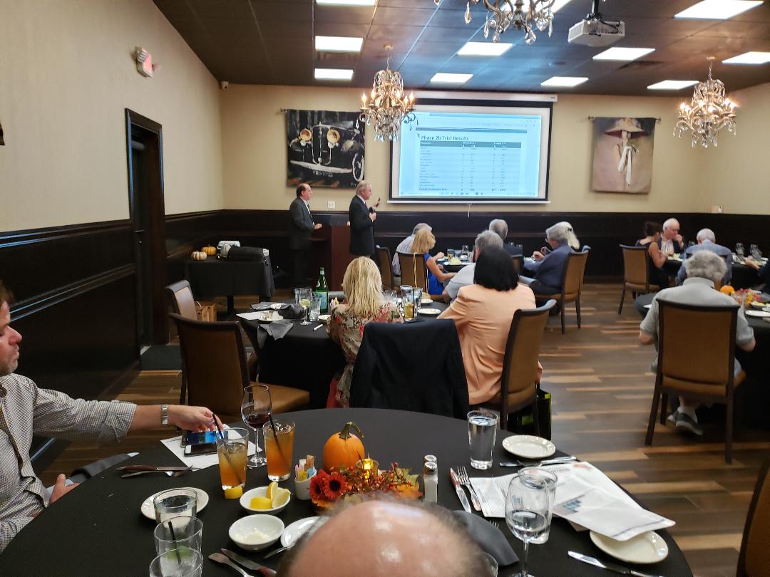 Guests seated at round tables watching a slide of clinical trial results