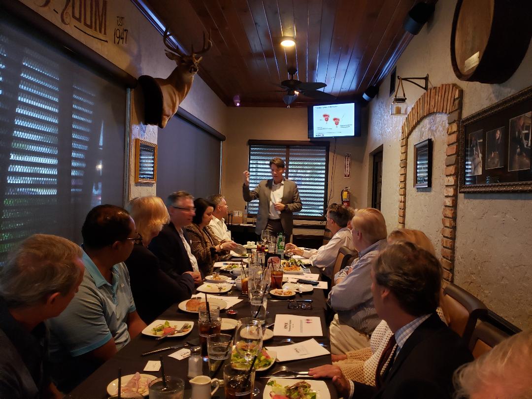 A presenter addressing guests along a long table beneath a wall-mounted screen