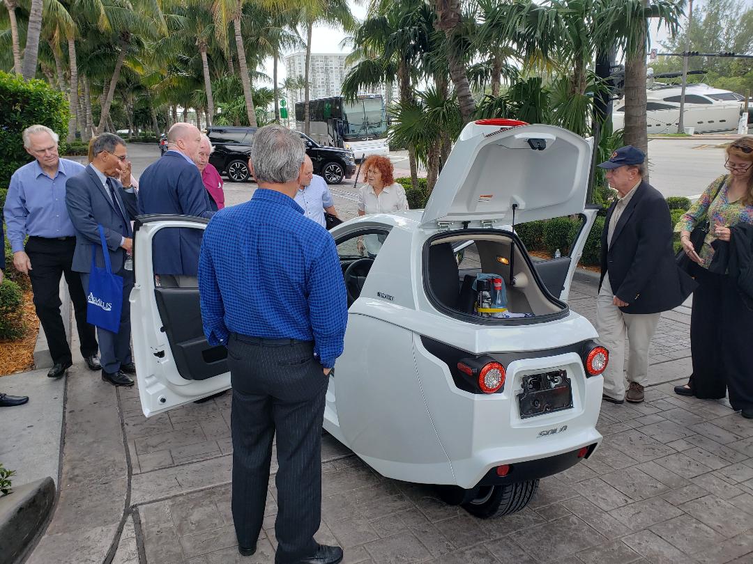 A group of attendees gathered around a white electric vehicle with doors open