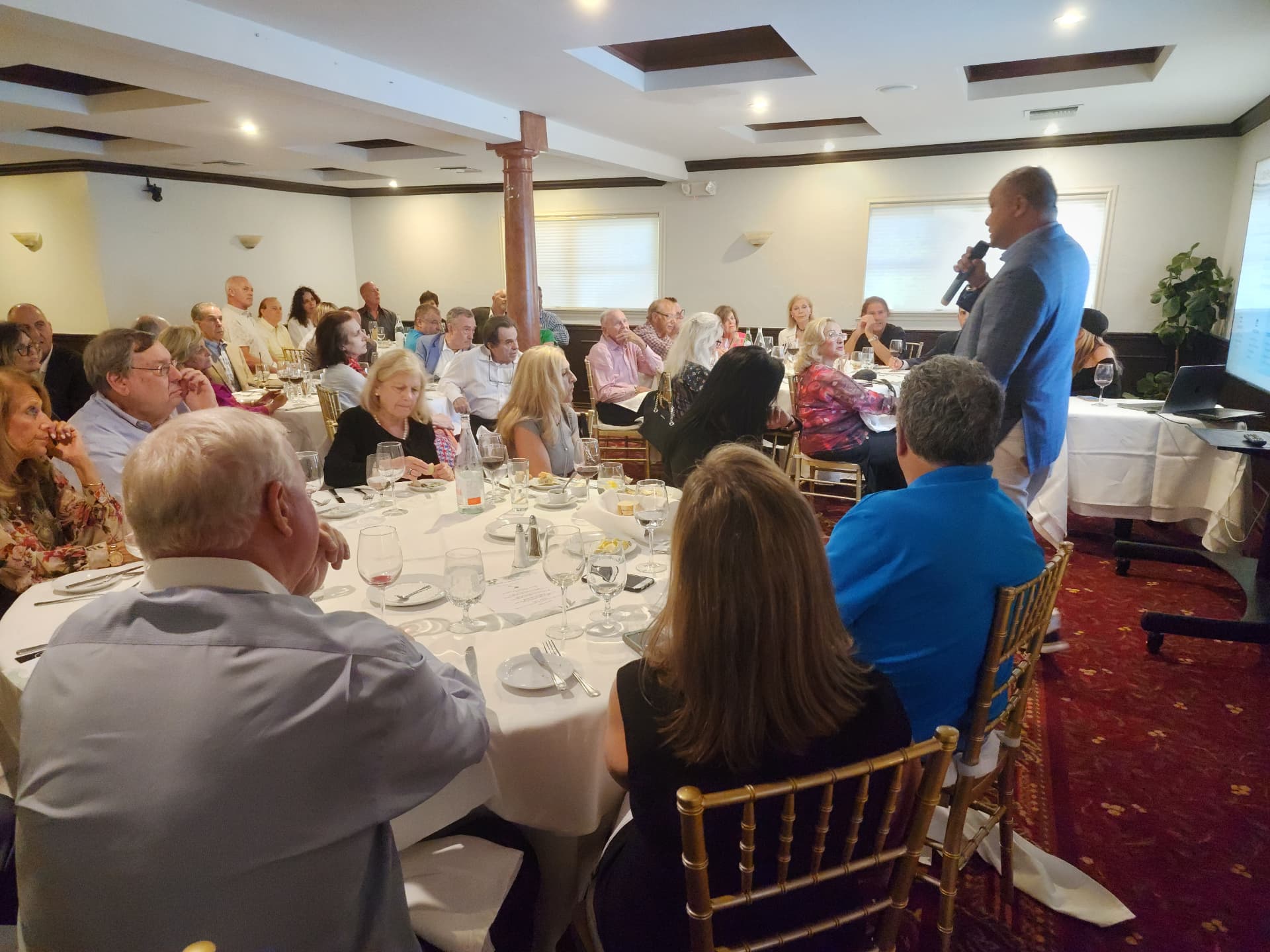 A host speaking with a microphone to guests seated at round tables in a banquet room