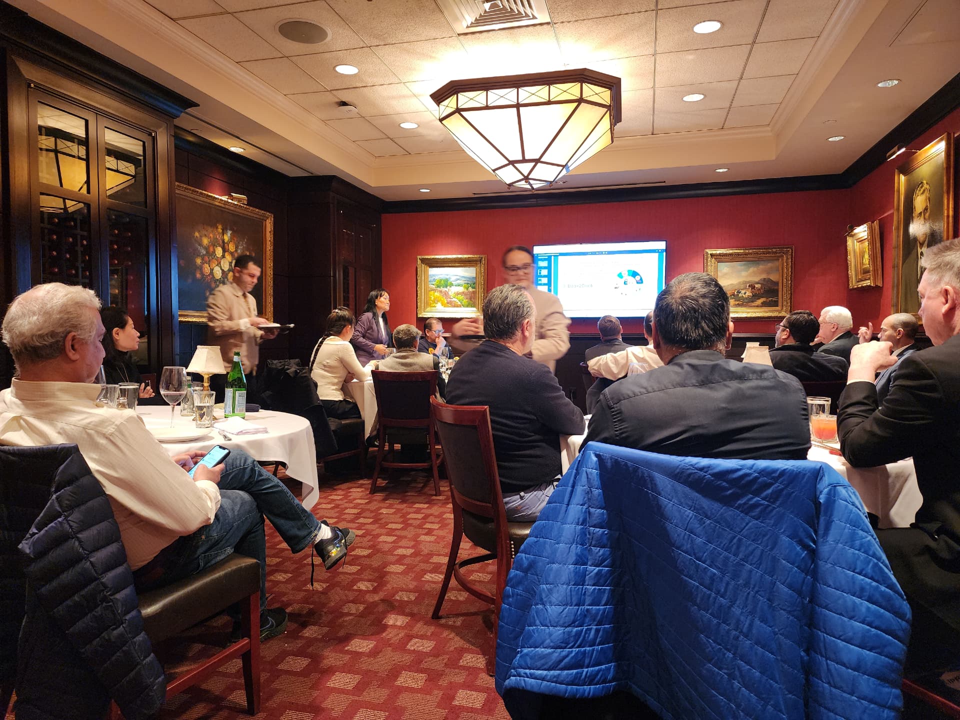 An analyst presenting a chart to guests seated at tables in a red dining room